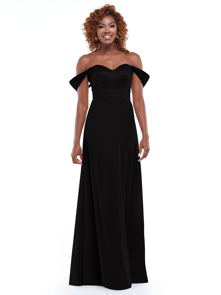 A woman with curly hair models Avery Austin’s 80164, an off-the-shoulder black dress with a delicate lace bodice. She stands smiling against a plain white background, arms relaxed by her sides.