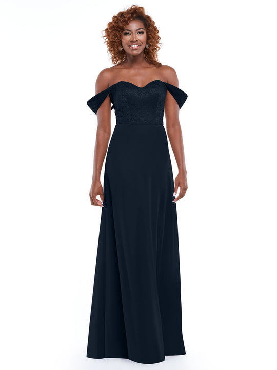 A smiling woman with curly hair models Avery Austin’s floor-length dress (style 80164) featuring an off-the-shoulder lace bodice and chiffon skirt, standing against a plain white background.
