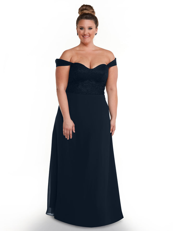 A woman with her hair in a bun is smiling and wearing the Avery Austin TH-80164 dress, featuring an off-the-shoulder lace bodice and floor-length dark navy chiffon skirt, standing against a plain white background.