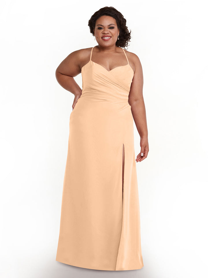 A woman smiles against a plain white background wearing Avery Austin style 80167, a blush floor-length chiffon bridesmaid dress with ruching, spaghetti straps, a sweetheart neckline, and a thigh-high slit.