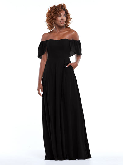 A woman with curly hair models Avery Austin's 80168—a black chiffon off-the-shoulder A-line dress with pockets and short ruffled sleeves. She stands against a white background, smiling slightly with one hand in her pocket.