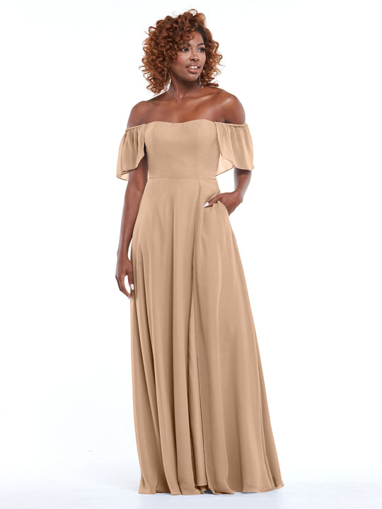 A woman wears Avery Austin’s 80168 chiffon off-the-shoulder A-line dress in Buff with pockets and short flutter sleeves, standing against a white background. She has curly hair, smiles slightly, and has one hand in her pocket.
