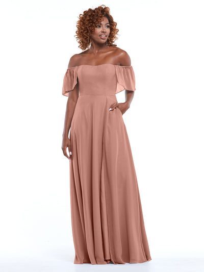 A woman with curly hair models Avery Austin’s 80168: a chiffon, off-the-shoulder, floor-length dusty rose A-line dress with short flutter sleeves and a flowing skirt, standing against a plain white background.