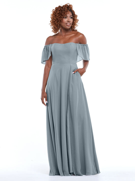 A woman with curly hair models Avery Austin’s 80168, a dusty blue chiffon off-the-shoulder A-line dress with pockets and flutter sleeves, against a plain white background.