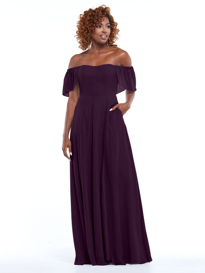 A woman, with curly hair and one hand in her pocket, models the Avery Austin 80168: a dark purple chiffon off-the-shoulder A-line dress featuring short ruffled sleeves and convenient pockets, standing against a white background.