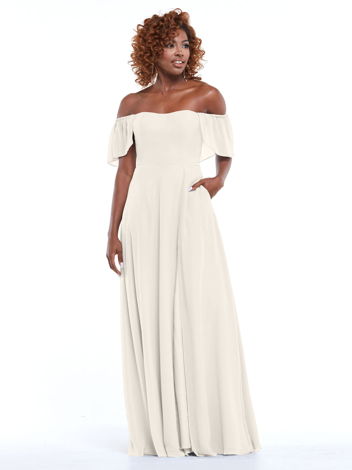 A woman with curly hair models Avery Austin’s 80168: a chiffon, off-the-shoulder, floor-length light cream A-line dress with short flutter sleeves and fitted bodice, standing against a plain white background.