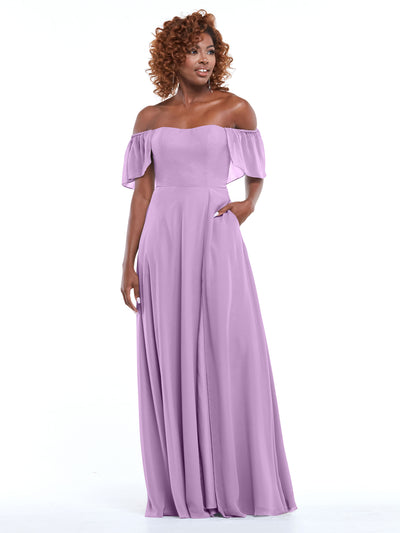A woman with curly hair poses in Avery Austin style 80168, a lavender A-line chiffon gown with off-the-shoulder ruffled sleeves and pockets, standing against a white background with one hand in her pocket.