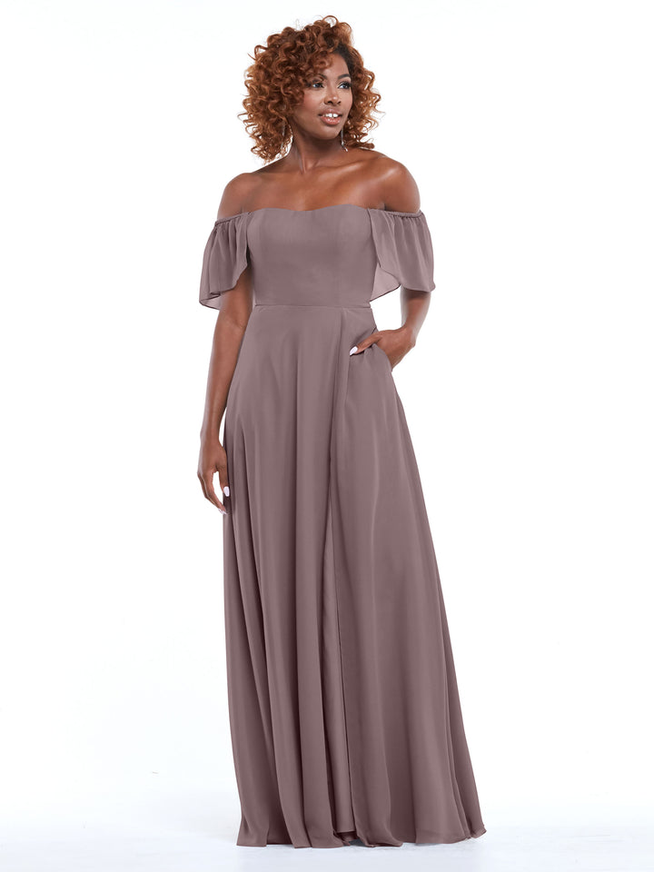 A woman models Avery Austin’s 80168 mauve chiffon off-the-shoulder, floor-length A-line dress with short ruffled sleeves, standing against a white background with her hand in the pocket.