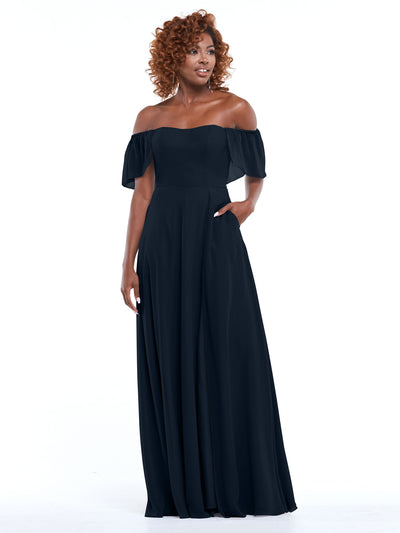 A woman with curly hair models the Avery Austin 80168, a floor-length black chiffon off-the-shoulder A-line dress with pockets and short sleeves, standing against a white background.