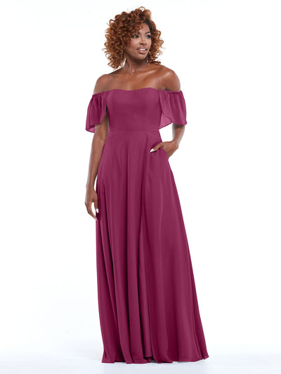 A woman wears Avery Austin 80168, a magenta chiffon off-the-shoulder A-line gown with ruffled sleeves and pockets. She stands against a white background, smiling with curly hair and one hand in her pocket.