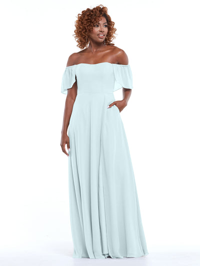 A woman in Avery Austin’s 80168 light blue chiffon off-the-shoulder A-line dress with pockets, ruffled short sleeves, and a flowing skirt stands against a white background, smiling and looking slightly to the side.