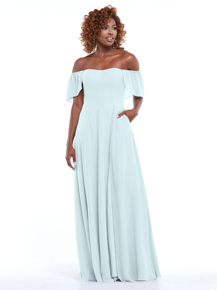 A woman in Avery Austin’s 80168 light blue chiffon off-the-shoulder A-line dress with pockets, ruffled short sleeves, and a flowing skirt stands against a white background, smiling and looking slightly to the side.