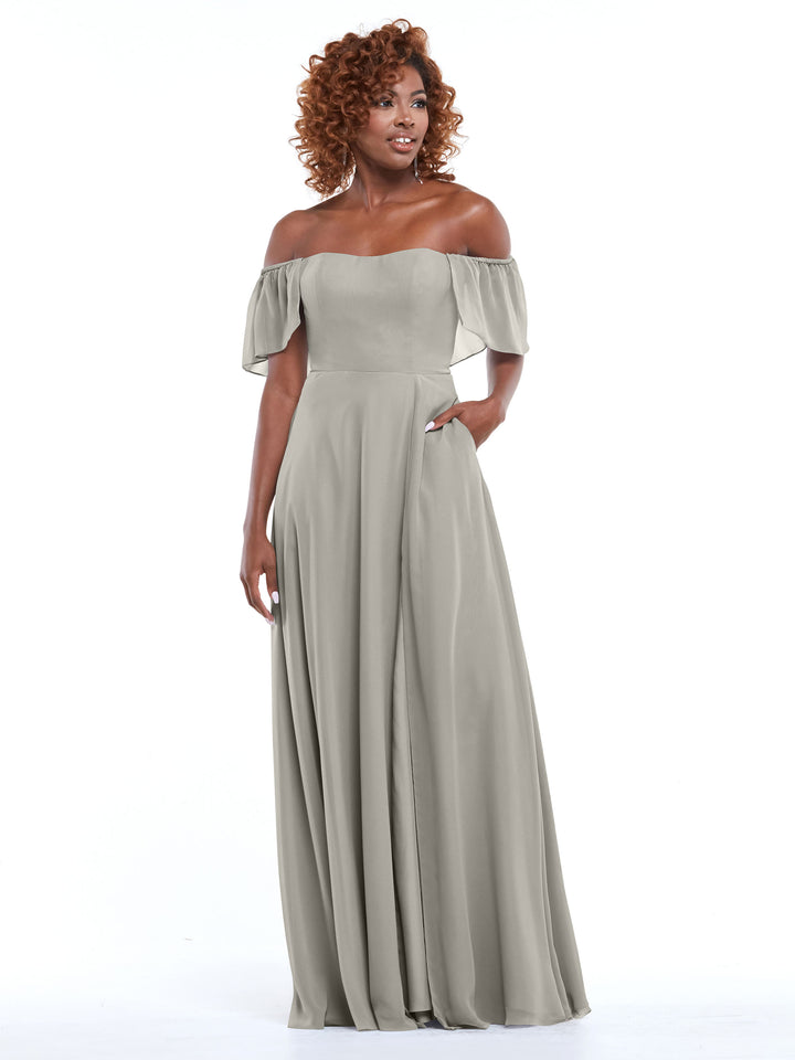 A woman with curly hair models Avery Austin’s 80168: a light gray, chiffon, off-the-shoulder, floor-length A-line dress with a fitted bodice and flowy skirt. She stands against a plain white background, smiling and looking to the side.