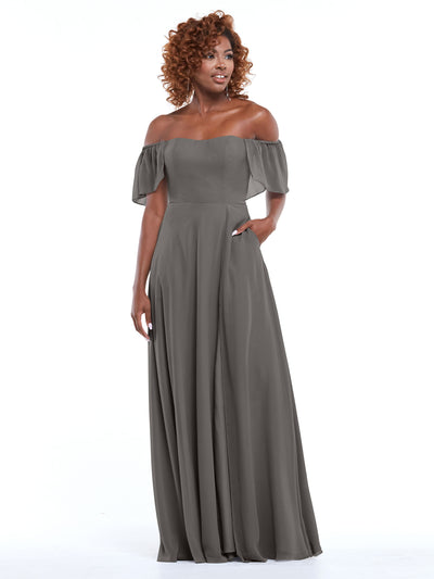 A woman with curly hair gently smiles while wearing the Avery Austin 80168: a gray, chiffon, off-the-shoulder A-line dress with pockets and short flowing sleeves, standing against a white background.