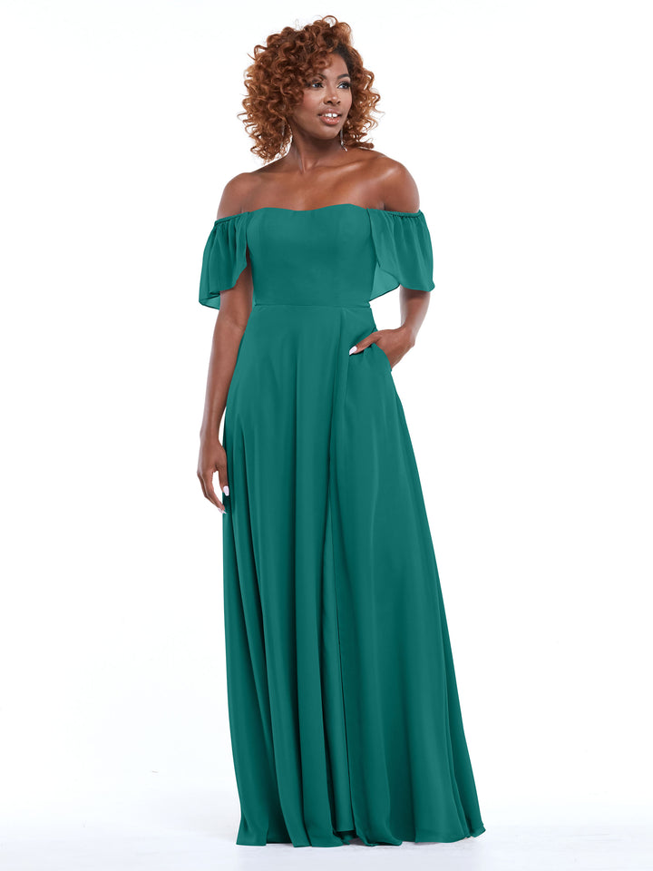 A woman with curly hair models Avery Austin’s 80168—a teal chiffon off-the-shoulder A-line dress with pockets and short sleeves—against a plain white background.