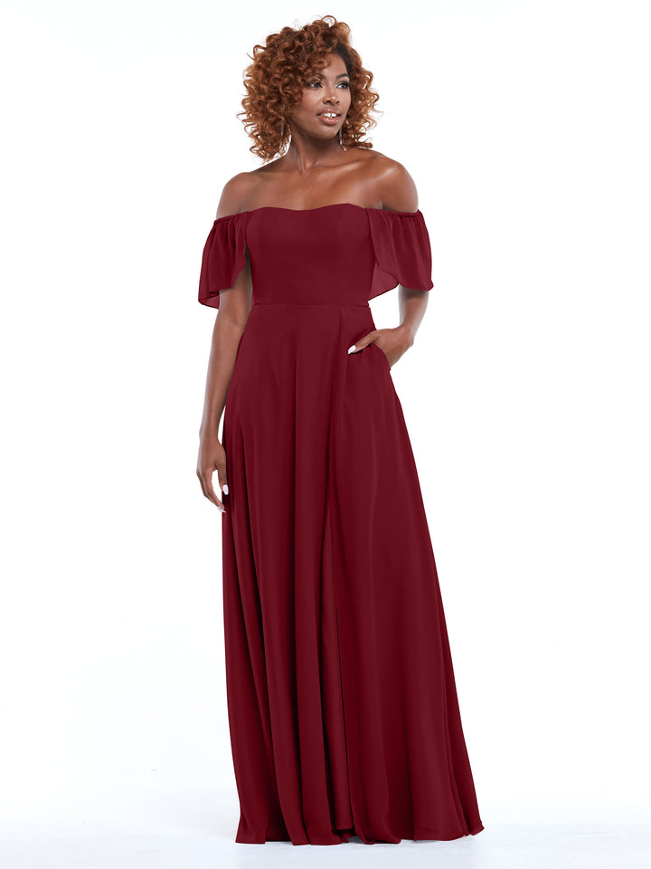 A woman in Avery Austin’s 80168 gown—a burgundy chiffon, off-the-shoulder, floor-length A-line dress with pockets and short ruffled sleeves—stands against a white background, smiling softly.