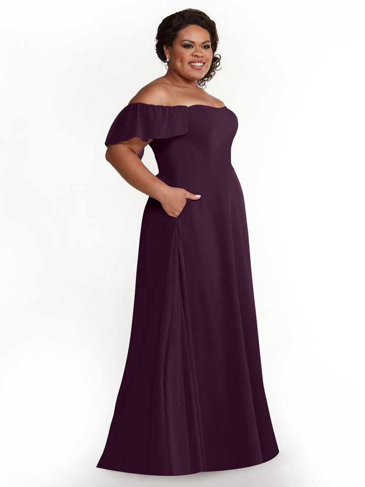 A smiling woman wears the Avery Austin 80168, an aubergine chiffon off-the-shoulder, floor-length A-line dress with flutter sleeves and side pockets, standing against a plain white background.