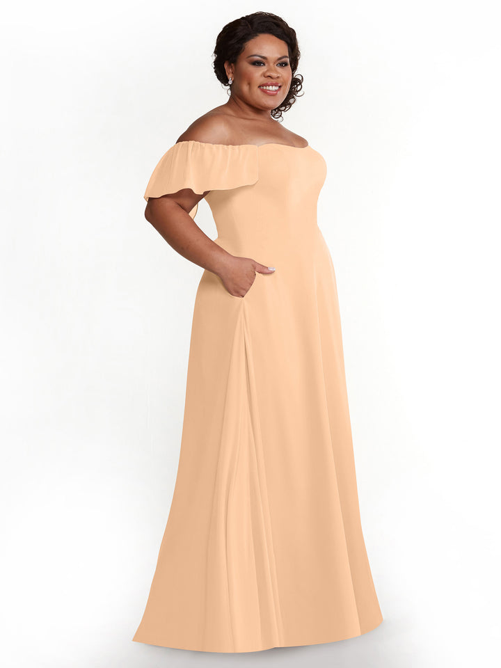 A woman smiles in the Avery Austin 80168 blush chiffon off-the-shoulder, floor-length A-line dress with short ruffled sleeves and side pockets, standing against a plain white background.