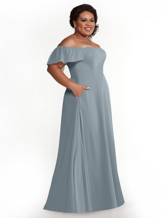 A woman smiles, wearing the Avery Austin 80168—a light blue chiffon off-the-shoulder A-line gown with pockets and short flutter sleeves—standing against a plain white background.