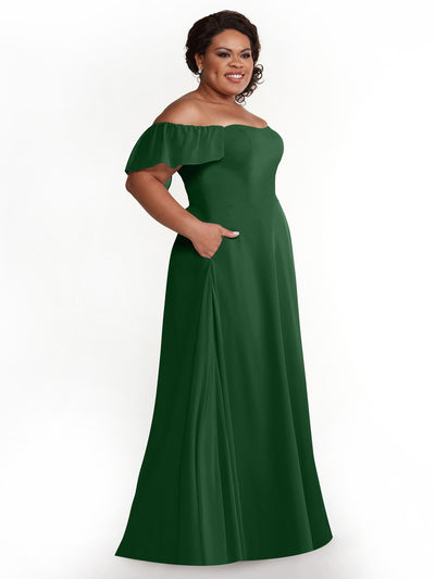 A woman smiles against a white background wearing Avery Austin style 80168, a chiffon off-the-shoulder emerald green A-line dress with flutter sleeves, floor length, and pockets.