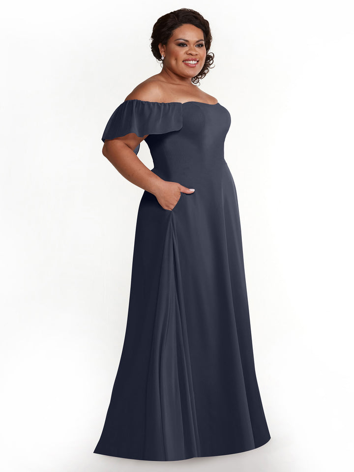 A woman stands smiling with her hands in the pockets of an Avery Austin 80168 dark blue chiffon off-the-shoulder A-line dress, set against a plain white background.