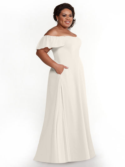 A woman stands smiling against a white background, wearing the Avery Austin 80168 cream chiffon off-the-shoulder A-line dress with short ruffled sleeves, floor-length skirt, and pockets.