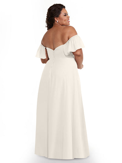 A woman in Avery Austin's 80168 cream chiffon off-the-shoulder A-Line dress with pockets stands facing away, smiling over her shoulder, her loose curls accented by pearl earrings.