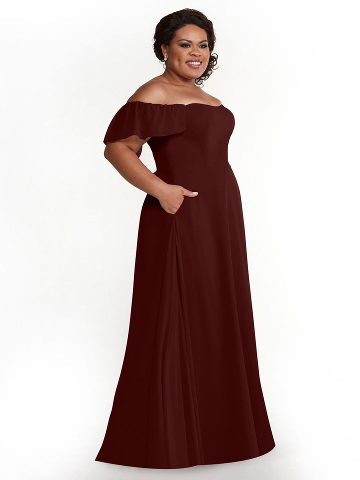 A woman with dark hair styled up smiles in an Avery Austin 80168 A-Line chiffon off-the-shoulder, floor-length Merlot gown with short flutter sleeves and pockets, standing against a plain white background.