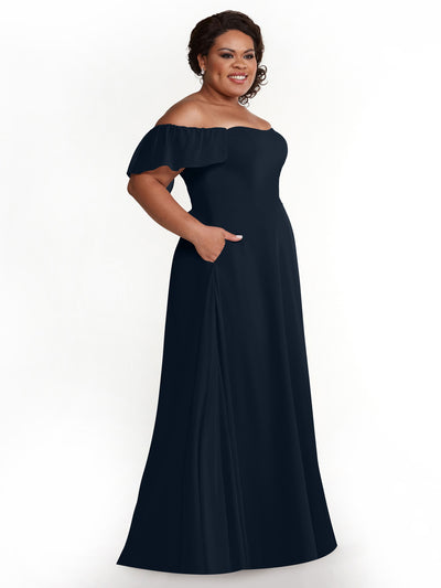 A woman smiles in the Avery Austin 80168 dress, a floor-length, dark navy chiffon A-line with off-the-shoulder ruffled short sleeves and pockets, posing against a plain white background.