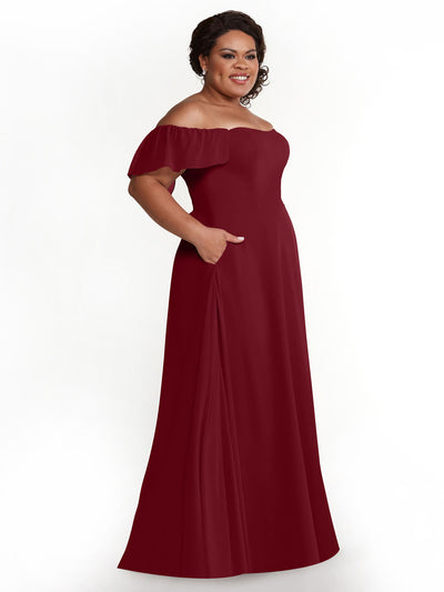 A smiling woman poses with her left hand in the pocket of Avery Austin’s 80168, a floor-length dark red off-the-shoulder chiffon A-line dress with pockets and short flutter sleeves, against a plain white background.