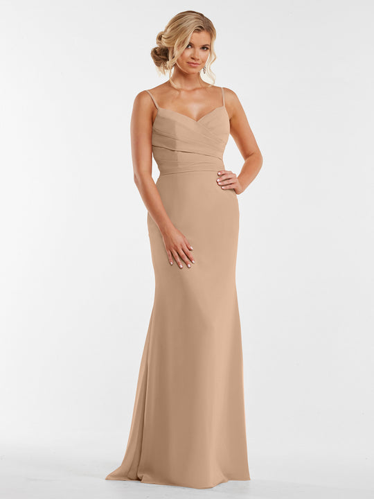 A woman poses with one hand on her hip, wearing the Avery Austin 80169—a buff floor-length fitted chiffon dress with thin straps and a pleated bodice—against a white background. Her hair is styled in a loose updo.