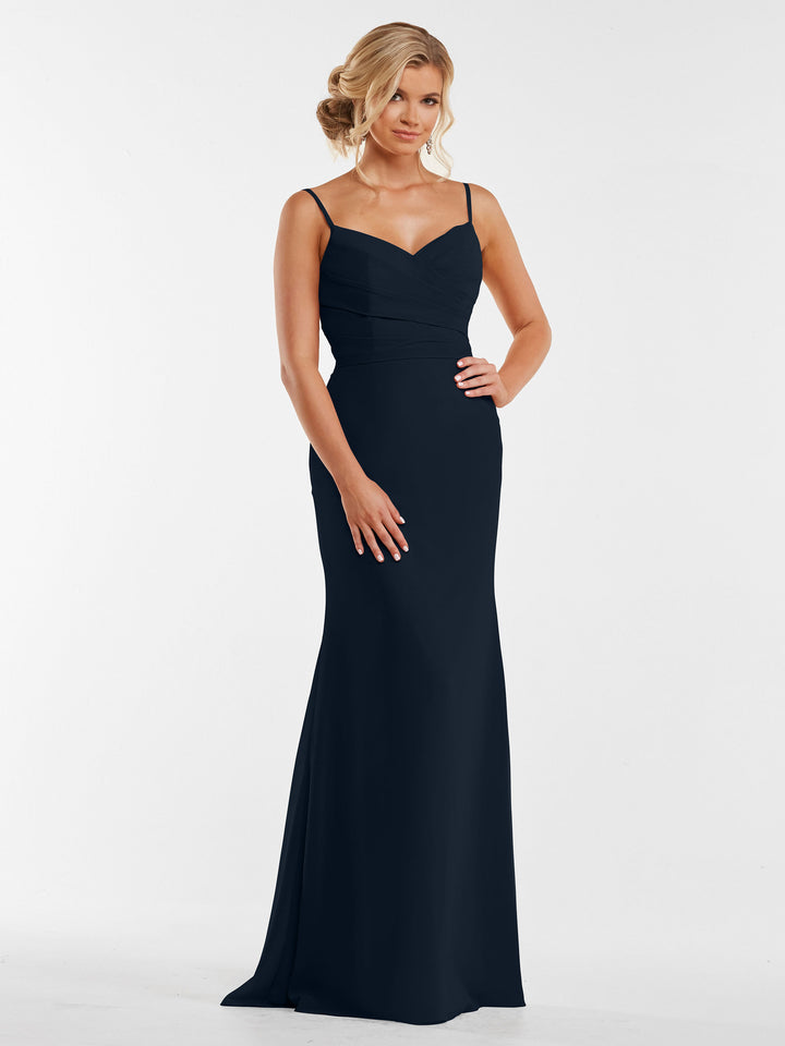 A woman with softly curled blonde hair wears Avery Austin style 80169, a fitted navy blue chiffon gown with spaghetti straps and a pleated bodice, standing against a plain white background with her hands resting in front.