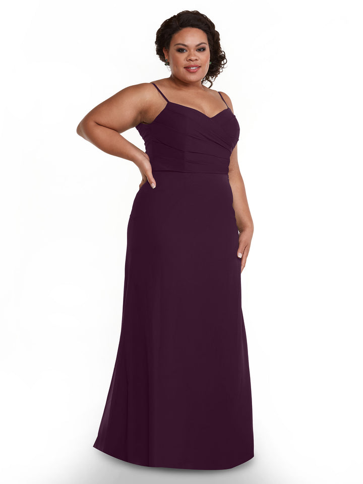A woman stands against a white background, smiling with one hand on her hip in Avery Austin’s 80169—a sleeveless, floor-length aubergine chiffon dress with thin straps and a pleated bodice.