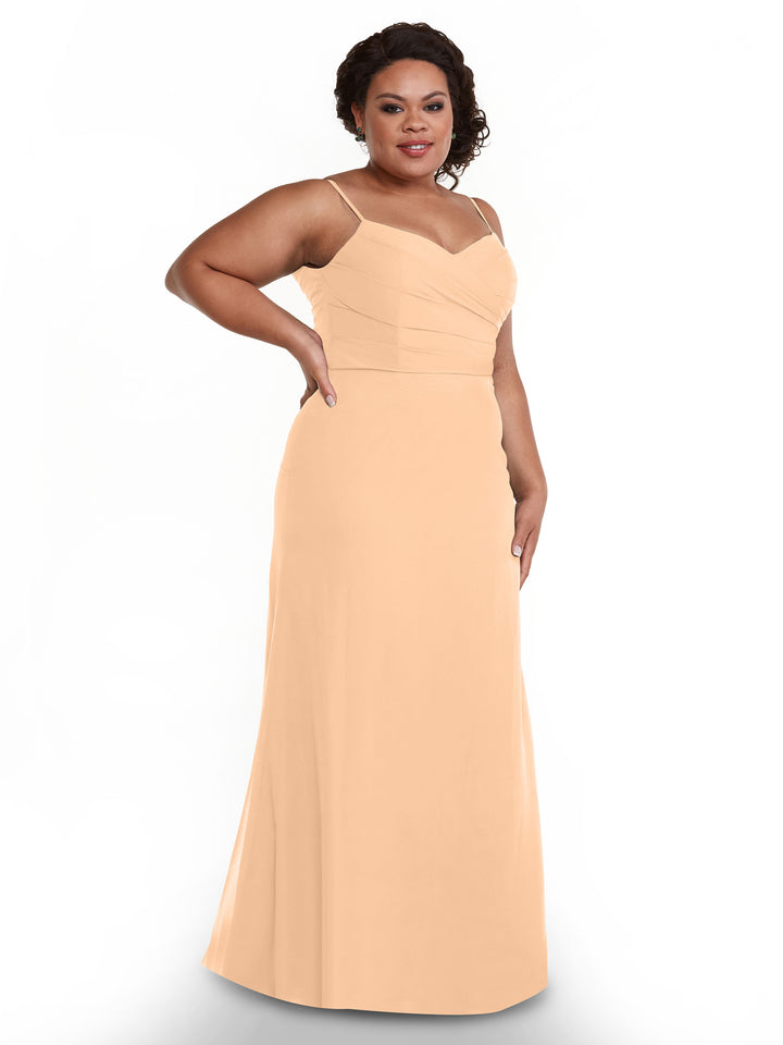 A woman poses confidently in Avery Austin’s blush chiffon dress (style 80169) featuring thin straps and a pleated bodice, set against a plain white background.