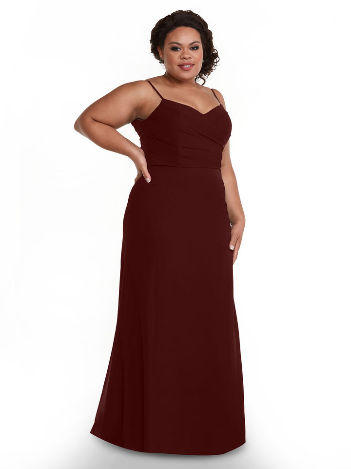 A woman stands against a plain white background, smiling with one hand on her hip while wearing the Avery Austin 80169 floor-length merlot chiffon dress with a pleated bodice and thin straps.