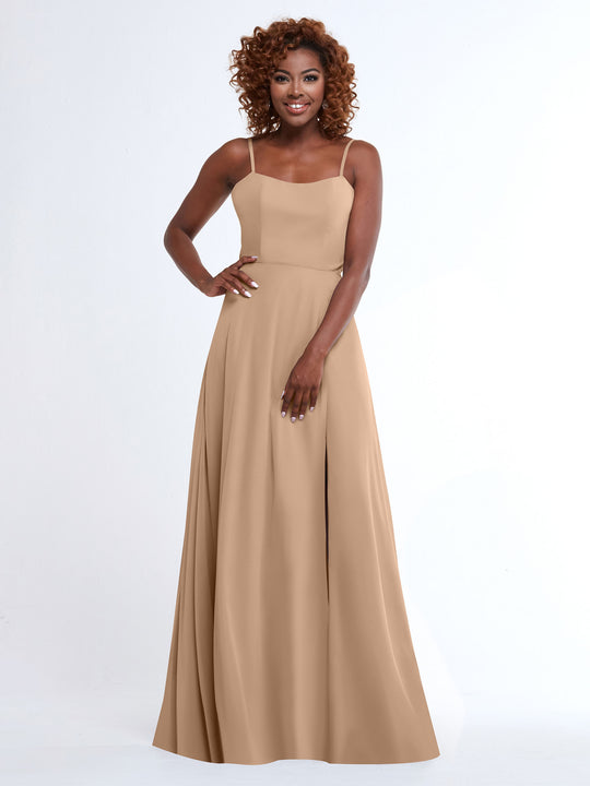A woman with curly hair smiles at the camera, wearing the Avery Austin 80170—a flowy, floor-length beige dress with spaghetti straps—against a plain white background.