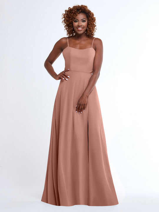 A woman with curly hair smiles while wearing Avery Austin’s 80170: a flowy, floor-length rose A-Line dress with spaghetti straps, standing against a plain white background.