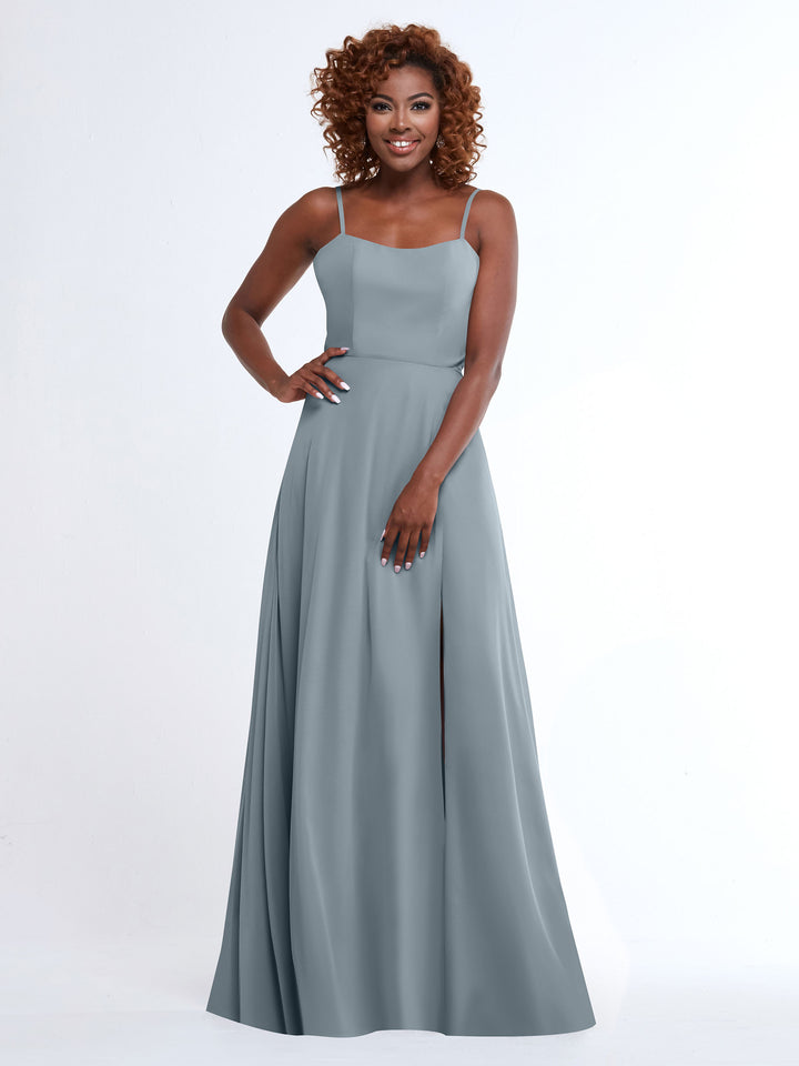 A woman with curly hair smiles while wearing the Avery Austin 80170, a flowy, floor-length light blue gown with spaghetti straps. She stands against a plain white background with one hand on her hip.