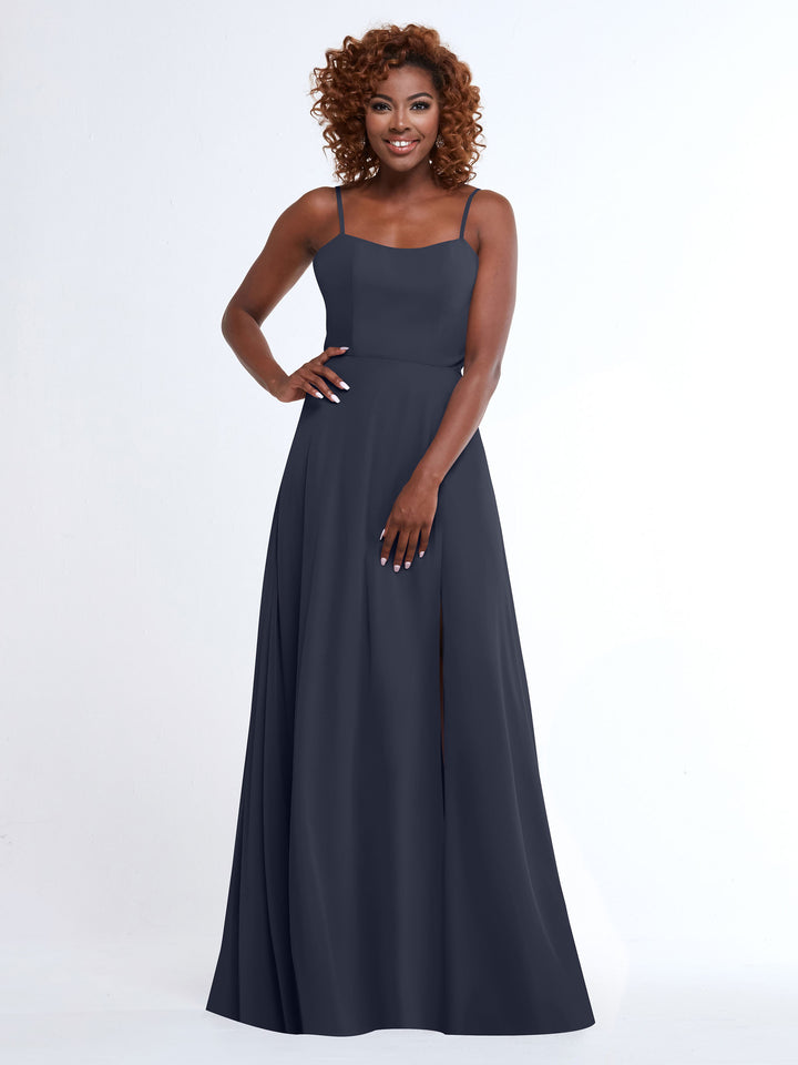 A woman with curly hair stands smiling in Avery Austin’s 80170, a sleeveless, floor-length dark blue A-line dress with spaghetti straps and a front slit, against a plain white background.