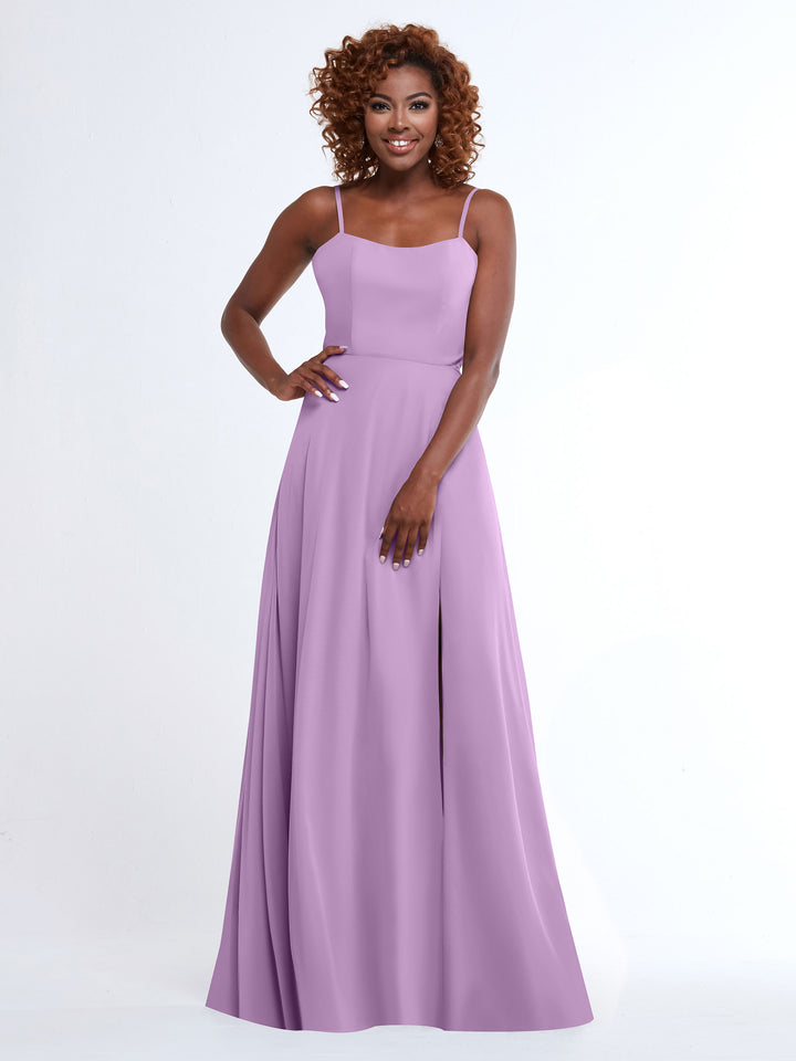 A woman with curly hair models Avery Austin’s 80170, a flowy, floor-length A-Line dress in lavender, standing against a white background and smiling with one hand on her hip.