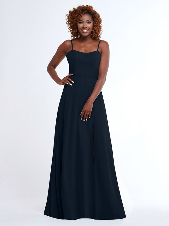 A woman with curly hair wears the Avery Austin 80170, a flowy navy blue A-Line dress with spaghetti straps and a side slit, standing against a white background and smiling at the camera.