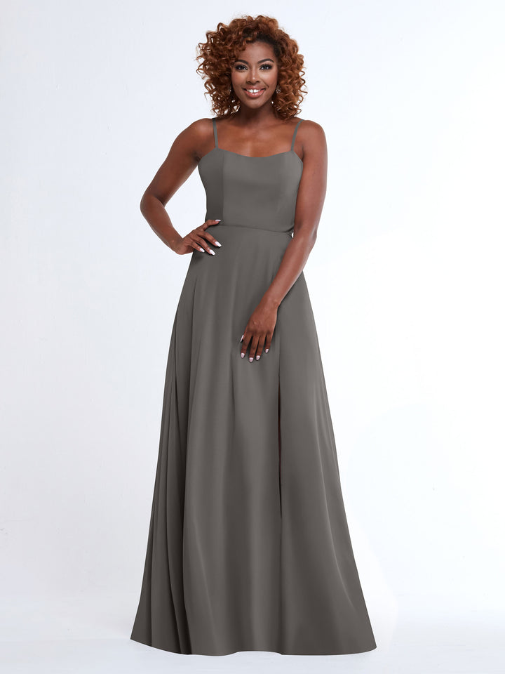 A woman with curly hair smiles while wearing the Avery Austin 80170, a sleeveless, floor-length grey A-line dress with spaghetti straps. She poses against a plain white background, one hand on her hip.