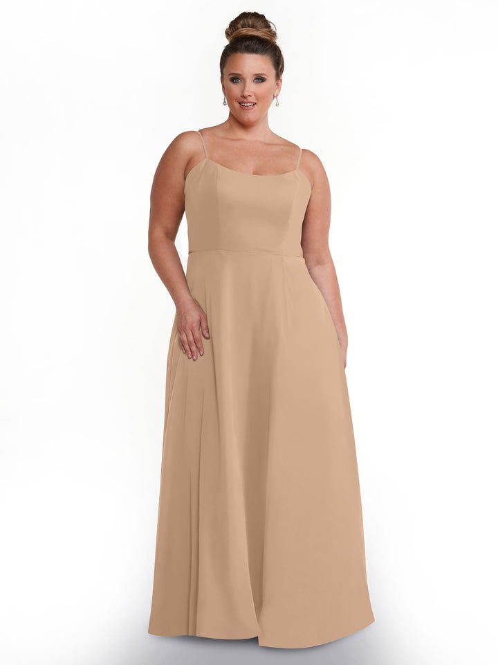 A woman wearing Avery Austin’s 80170 sleeveless floor-length light beige A-line dress with spaghetti straps stands against a white background, smiling slightly with her hair in an elegant bun.