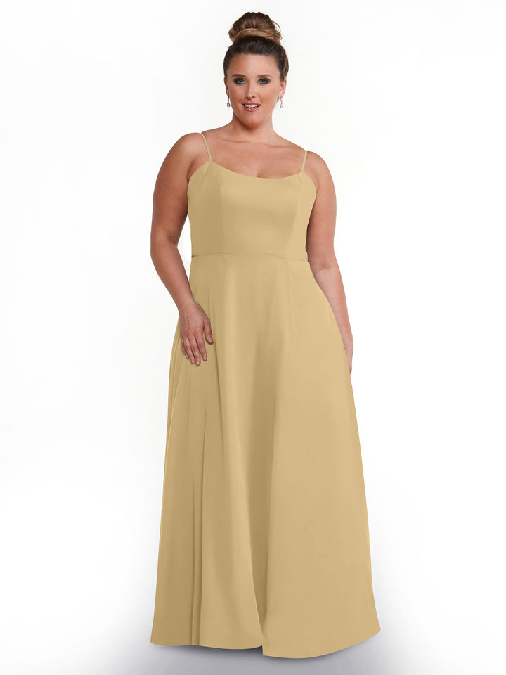 A woman poses against a white background wearing Avery Austin’s 80170—a long, light tan A-line dress with spaghetti straps. Her hair is styled in a neat bun, and she stands with one hand by her side and the other on her hip.