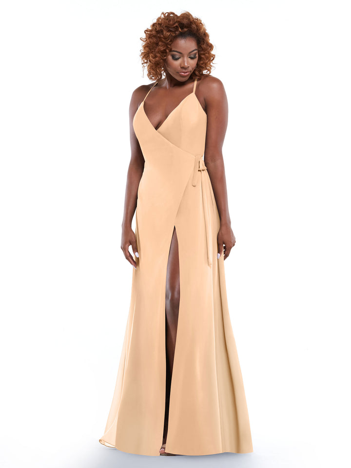 A woman with curly hair smiles and looks down while wearing the Avery Austin 80171—a long, sleeveless beige dress with a deep V-neck, thin straps, and a high slit—standing against a white background.