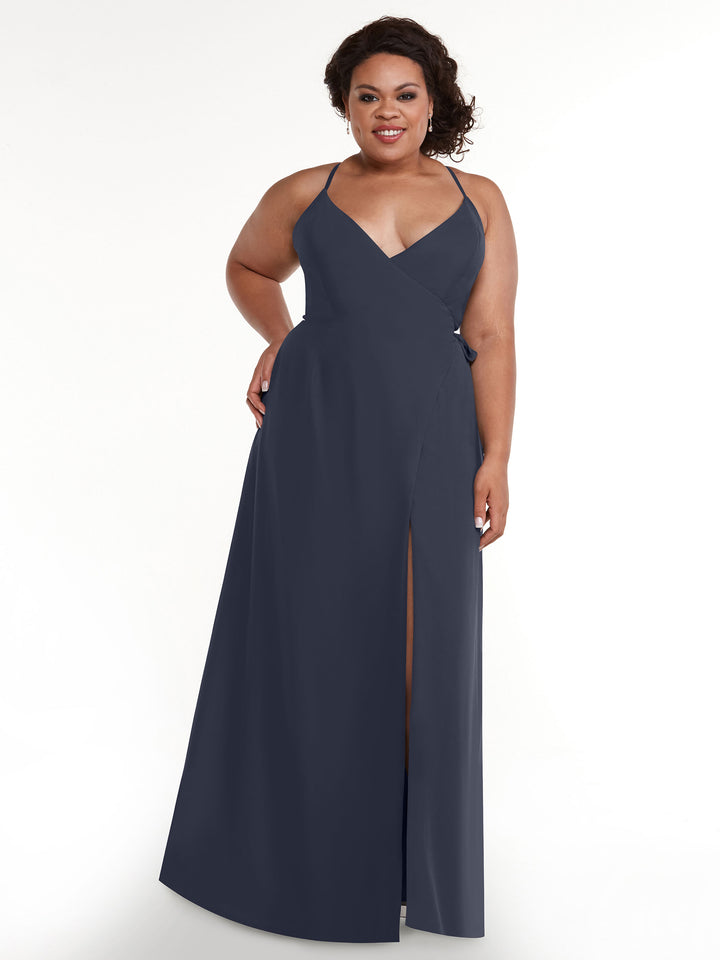 A woman poses with a smile and one hand on her hip, modeling Avery Austin’s floor-length, sleeveless navy slit dress (Style 80171) with a V-neckline against a plain white background.