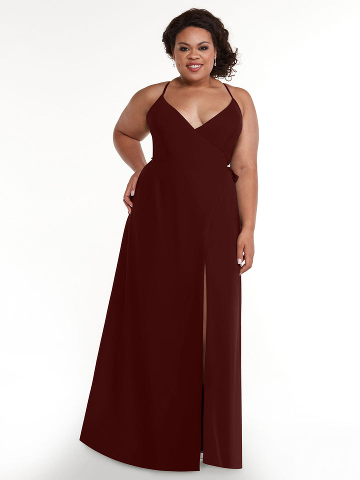 A woman with curly hair smiles in front of a white background, wearing Avery Austin’s 80171: a sleeveless, floor-length burgundy sheath dress featuring thin straps, a deep V-neckline, and a thigh-high slit.