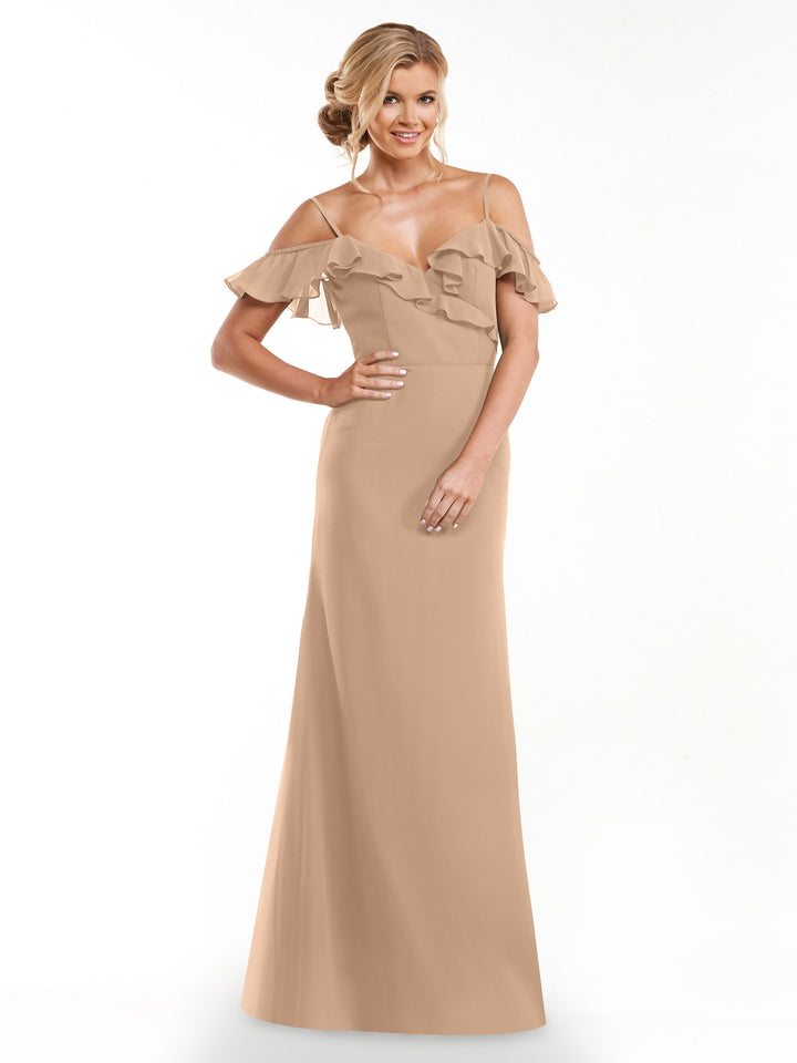 A woman stands against a white background, smiling with one hand on her hip and loose curls, wearing Avery Austin style 80172—a long, light beige A-line dress with a ruffled neckline and off-the-shoulder sleeves.