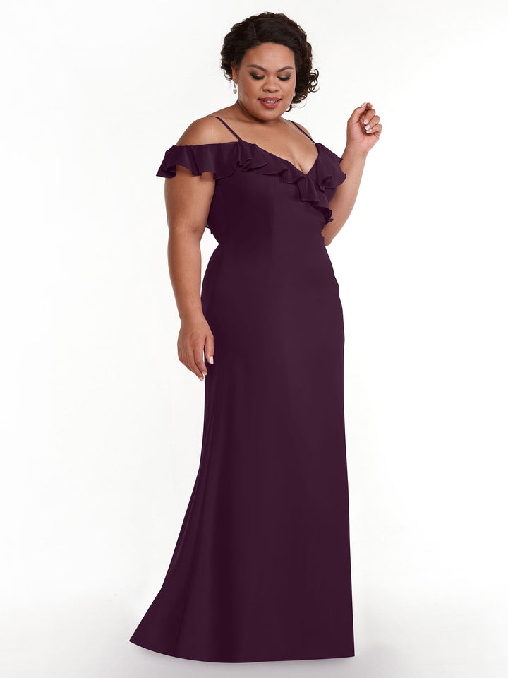 A woman models Avery Austin’s floor-length, deep purple A-Line dress (style 80172) with off-the-shoulder ruffled sleeves, standing against a white background and smiling gently while looking slightly down.