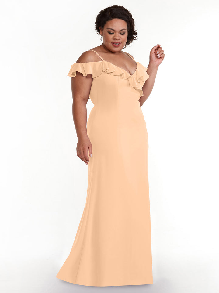 A woman wears Avery Austin’s 80172: a peach off-the-shoulder A-line dress with a ruffled neckline, standing against a white background and looking down gently.